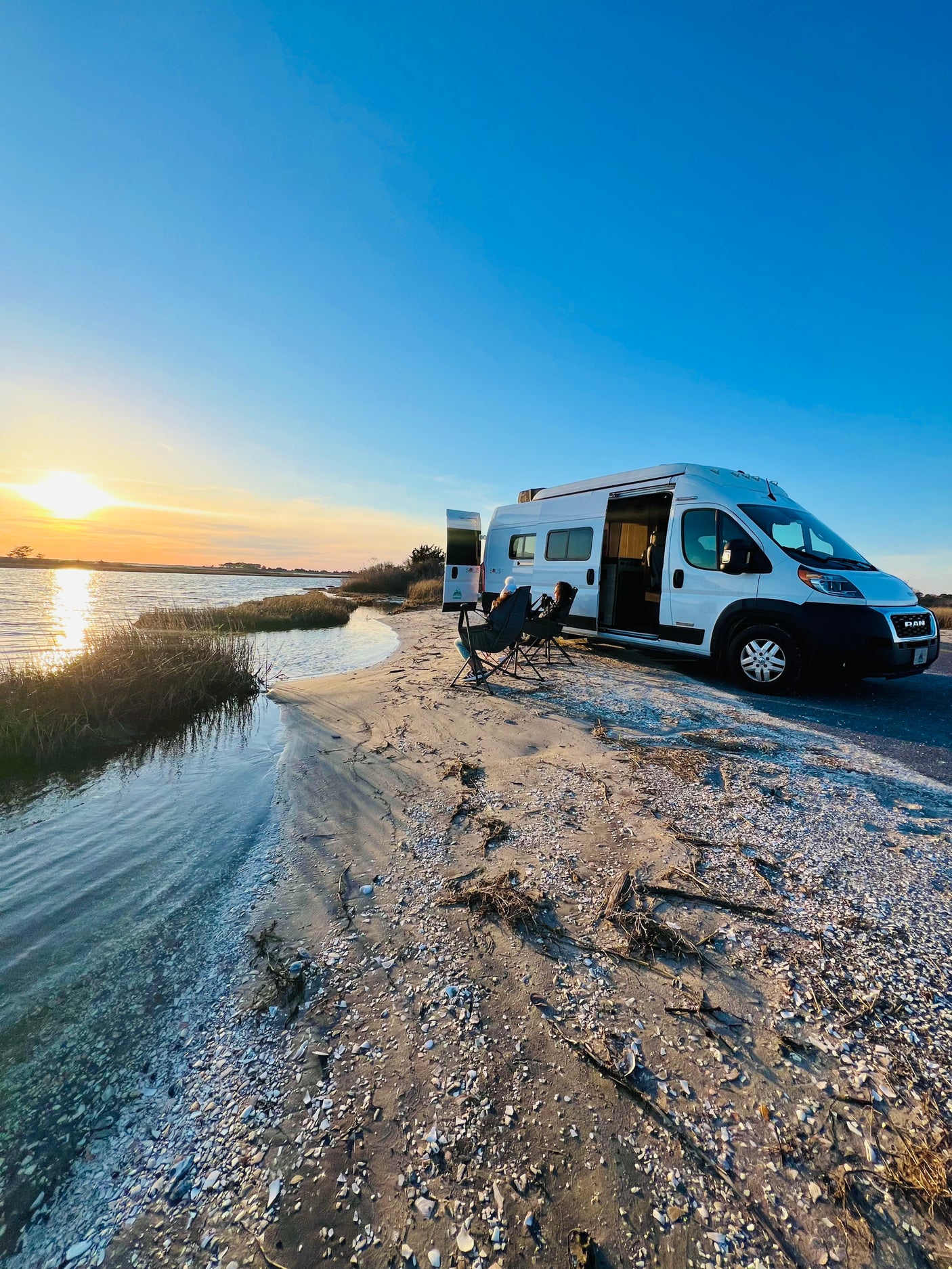 White rv rental by the water at sunset. Two people relax in chairs, enjoying the tranquil view. Ideal for outdoor adventures.