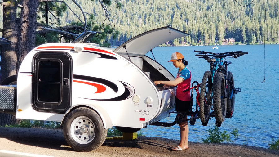 RV rental: a teardrop camper by a lake, with bikes ready for outdoor adventures. A person accesses the rear kitchen. Perfect camping setup!