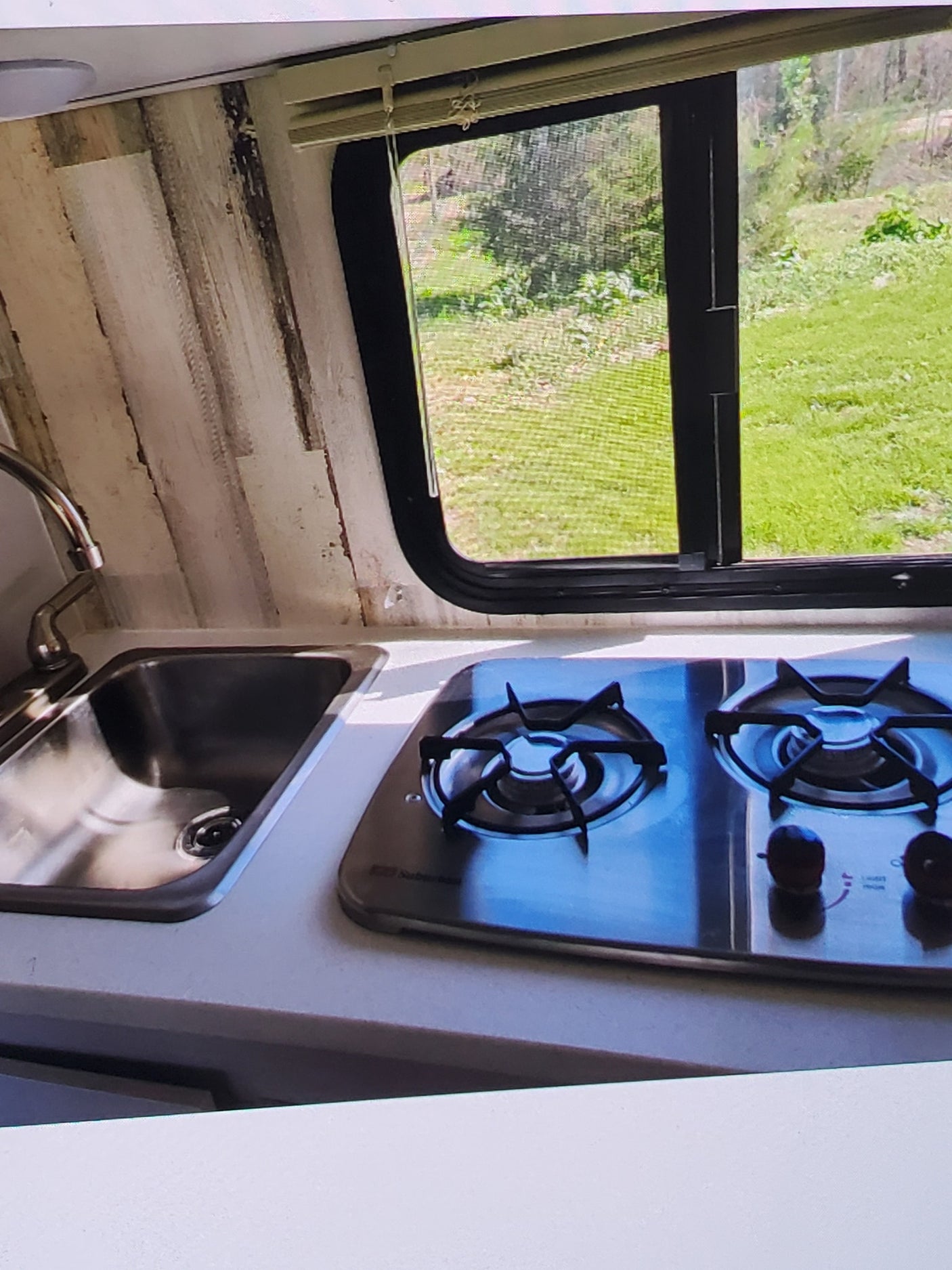 RV rental kitchen: Compact stainless steel sink and 2-burner stove, with a large window looking out to green nature.