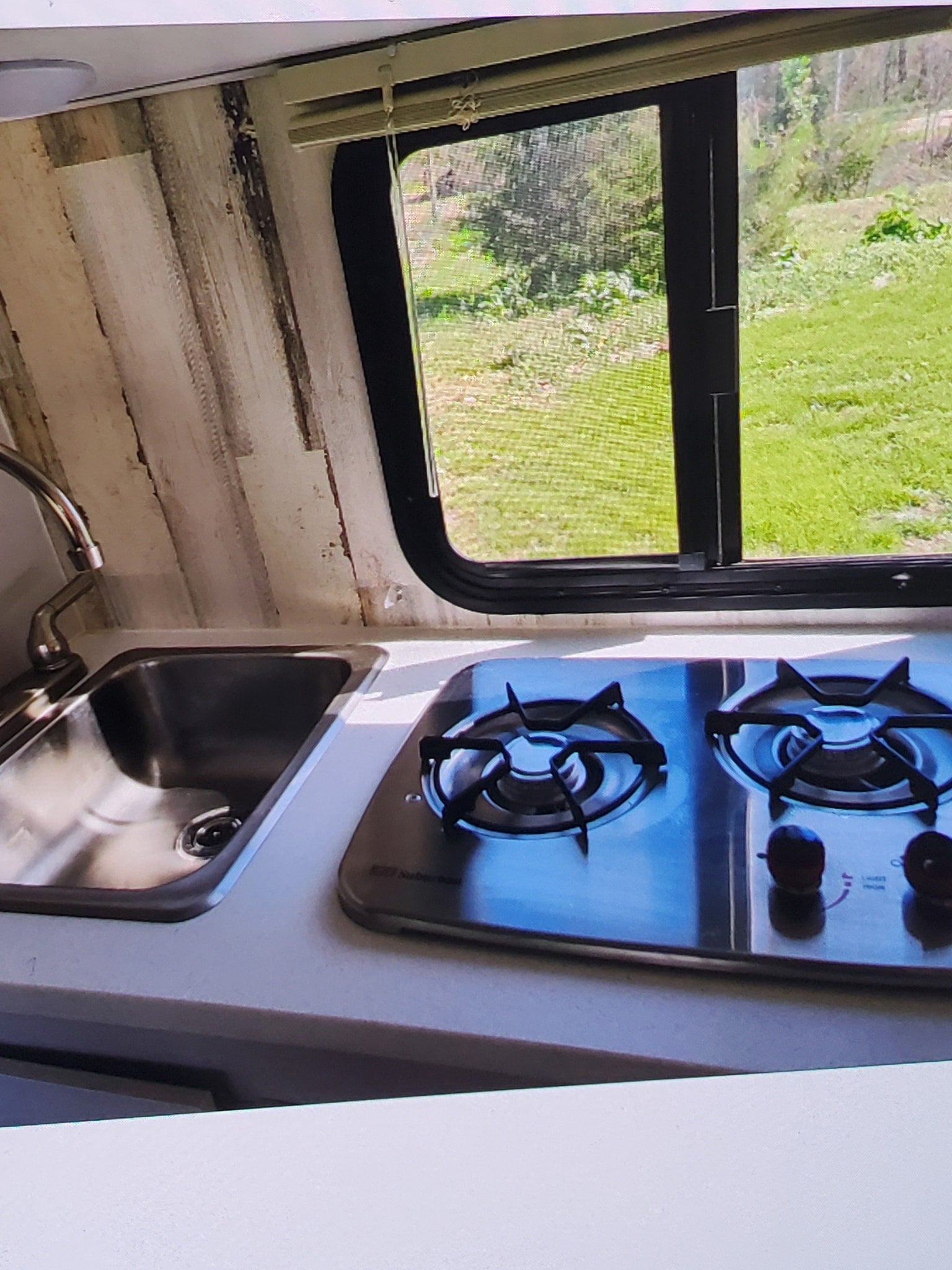 RV rental kitchen: Compact stainless steel sink and 2-burner stove, with a large window looking out to green nature.