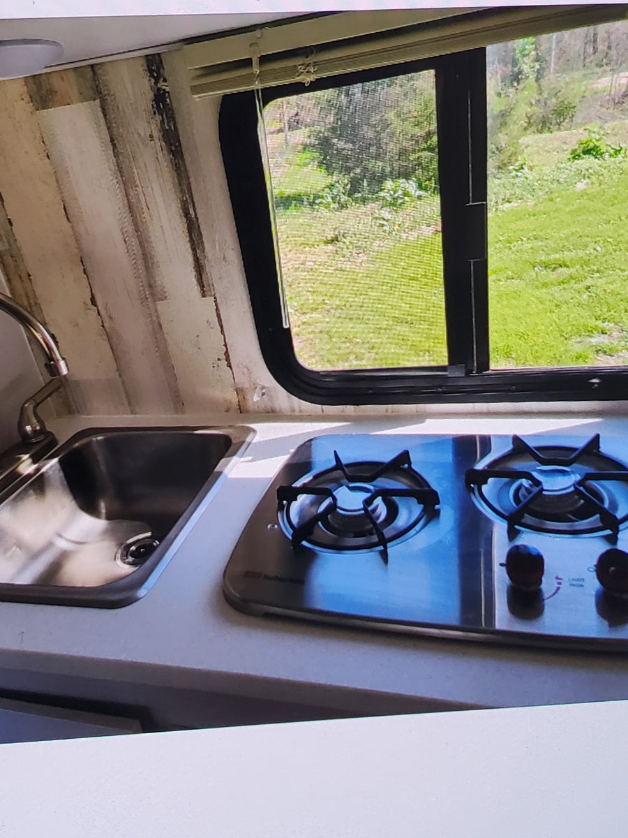 RV rental kitchen: Compact stainless steel sink and 2-burner stove, with a large window looking out to green nature.