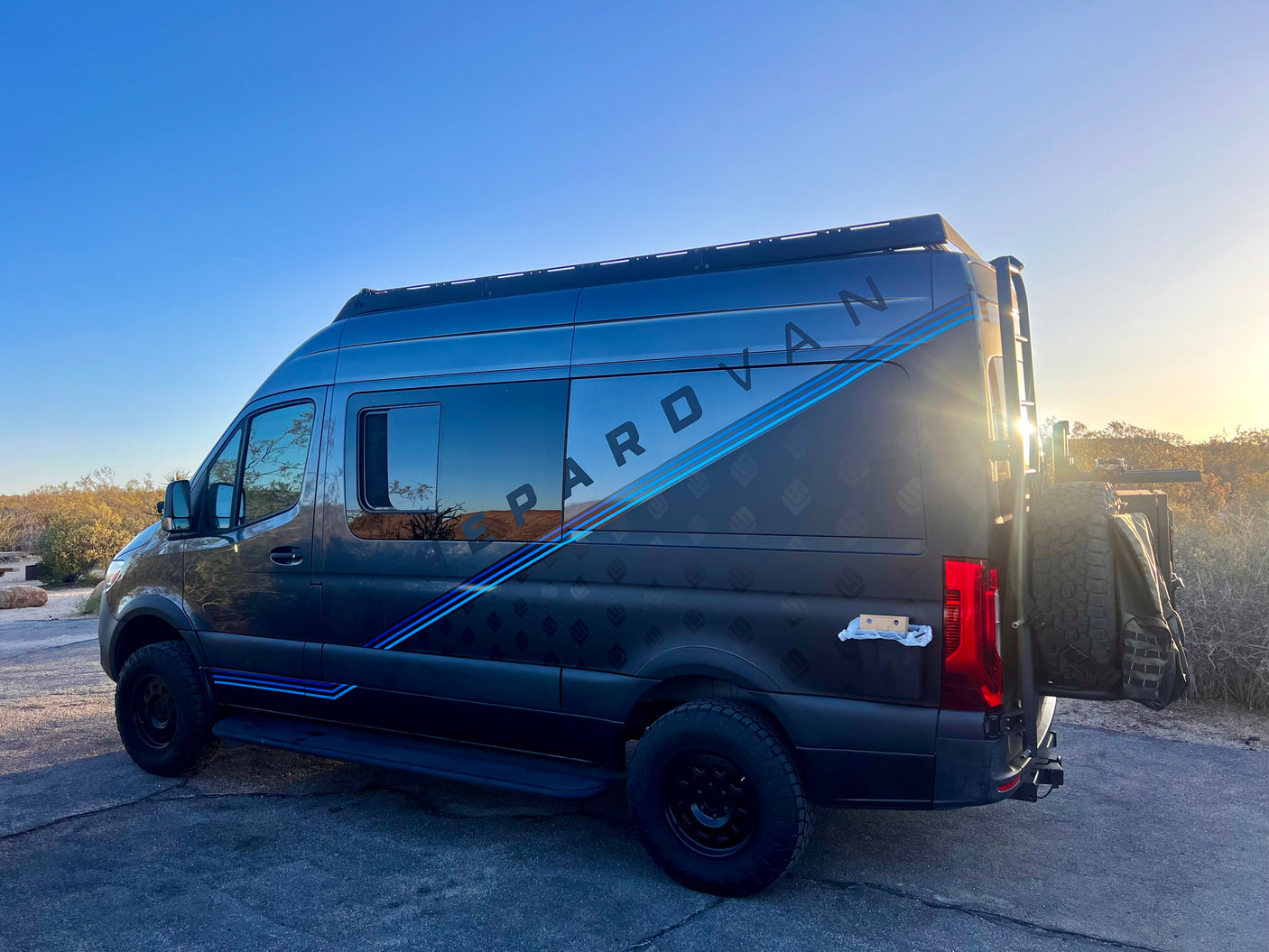 This dark gray "PAROVAN" Sprinter camper van, featuring a roof rack and rear tire, is ready for your RV rental adventure.