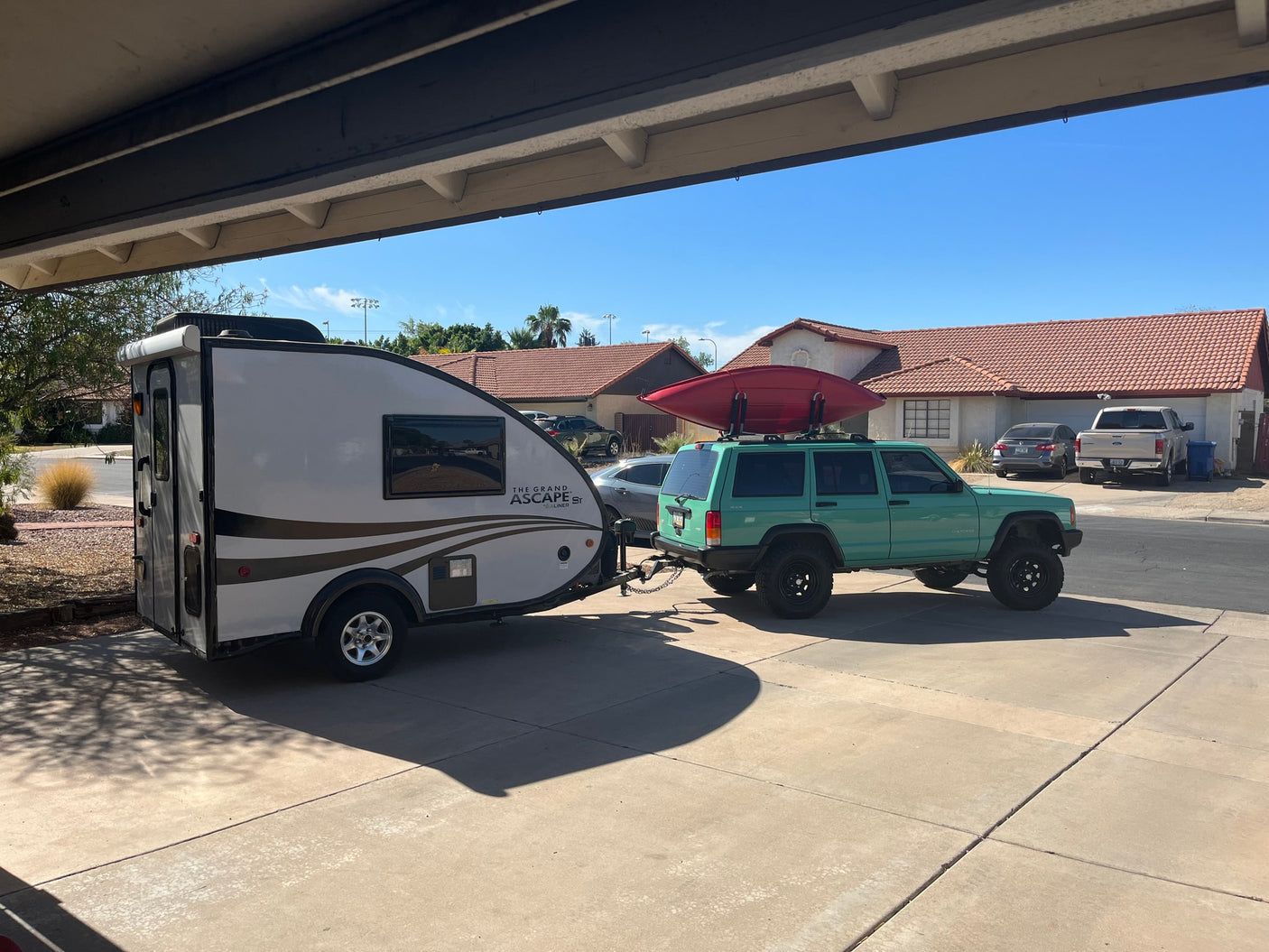 Teal Jeep with kayak, towing a white Grand Ascape travel trailer. This RV rental setup is parked in a sunny driveway.