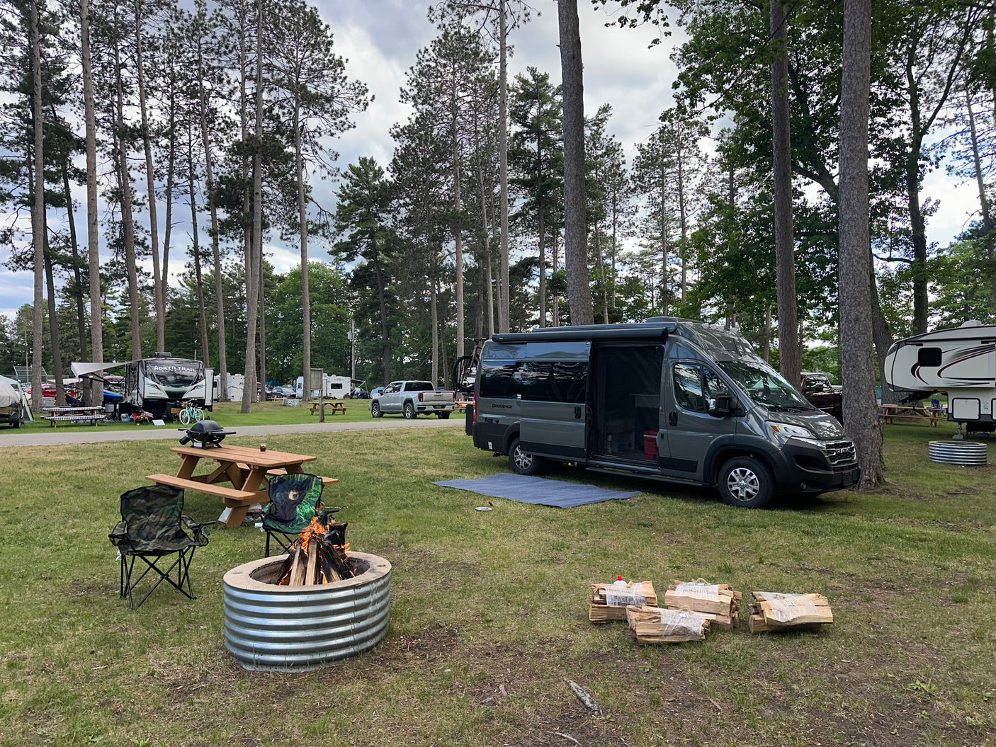 Peaceful campground scene with a grey camper van, blazing fire pit, and tall pines, ideal for an RV rental getaway.