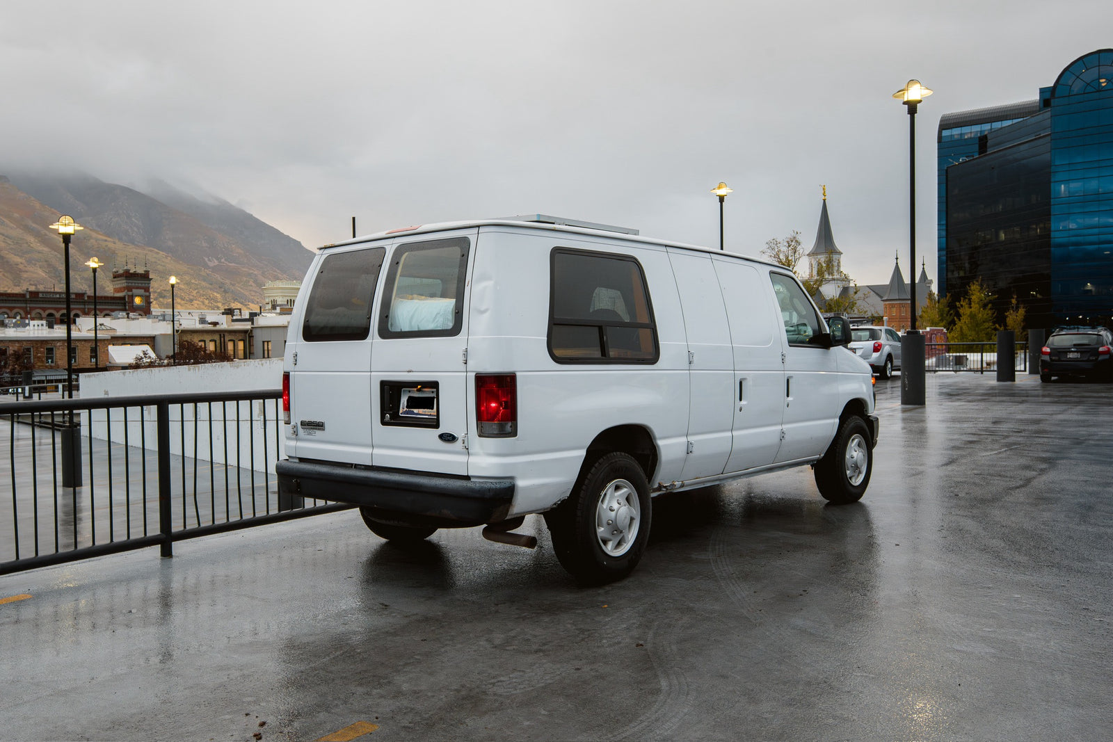 Explore with an rv rental! White van parked on wet street with city buildings, cloudy mountains, and streetlights.