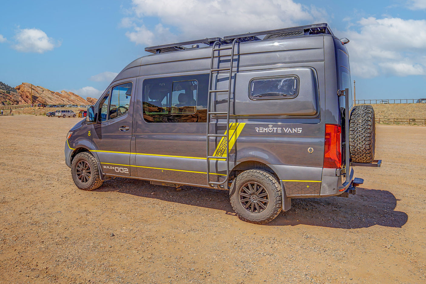 Adventure-ready gray Mercedes Sprinter RV rental van with off-road tires parked on a dirt lot near red rock formations.