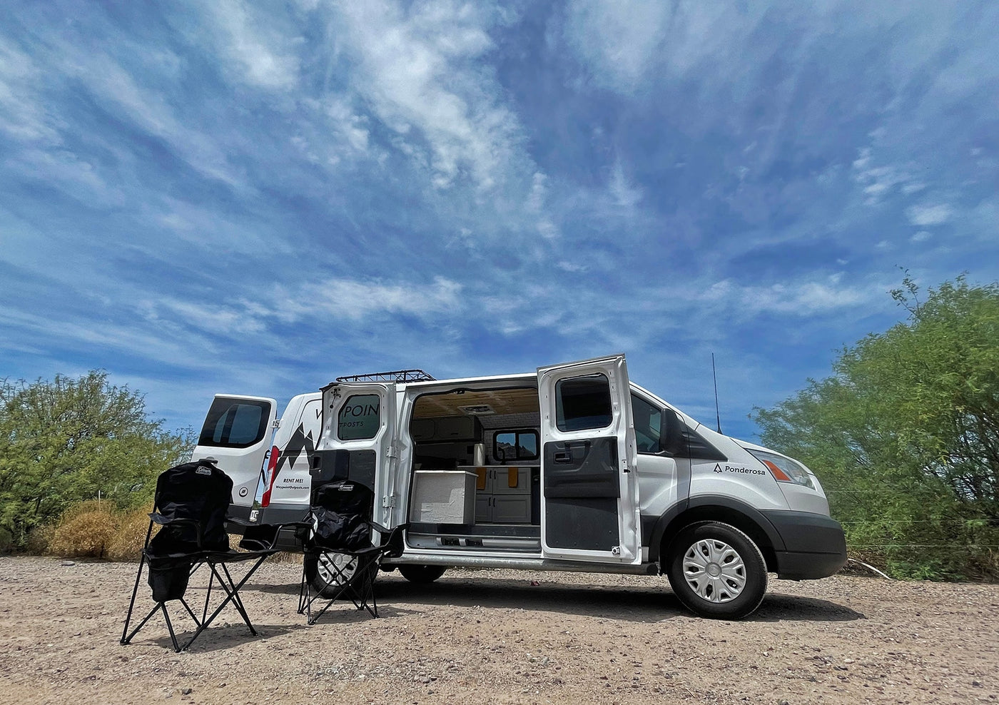 White RV rental van parked on a dirt road, side door open revealing a kitchenette, with two folding chairs set outside. — photo 9