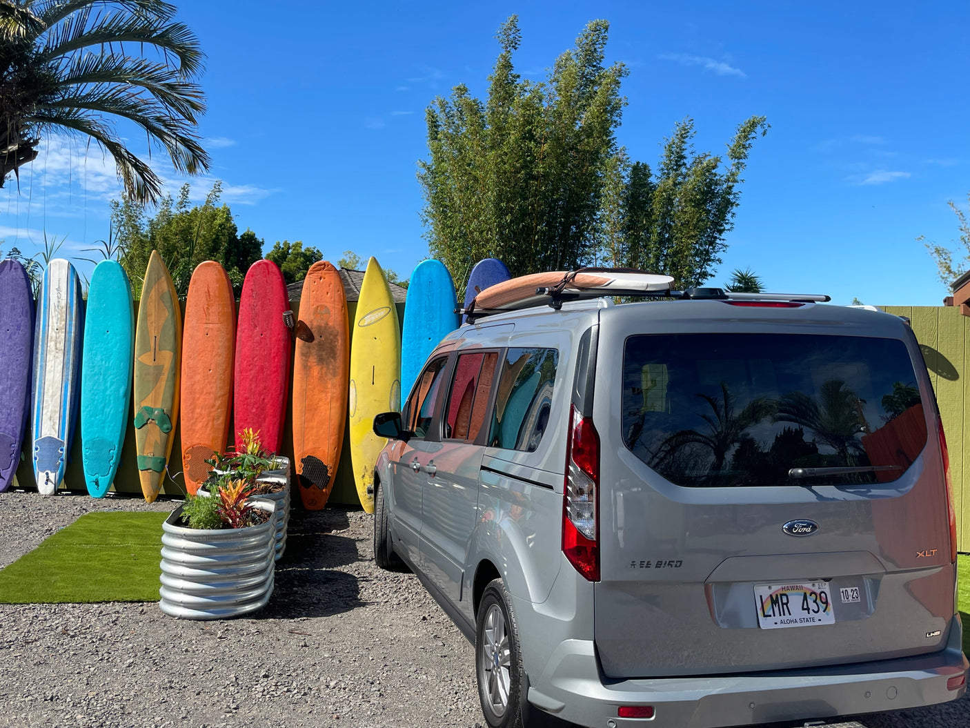Gray Ford van, an RV rental, with surfboards on its roof, parked by a fence lined with colorful surfboards under a blue Hawaii sky.