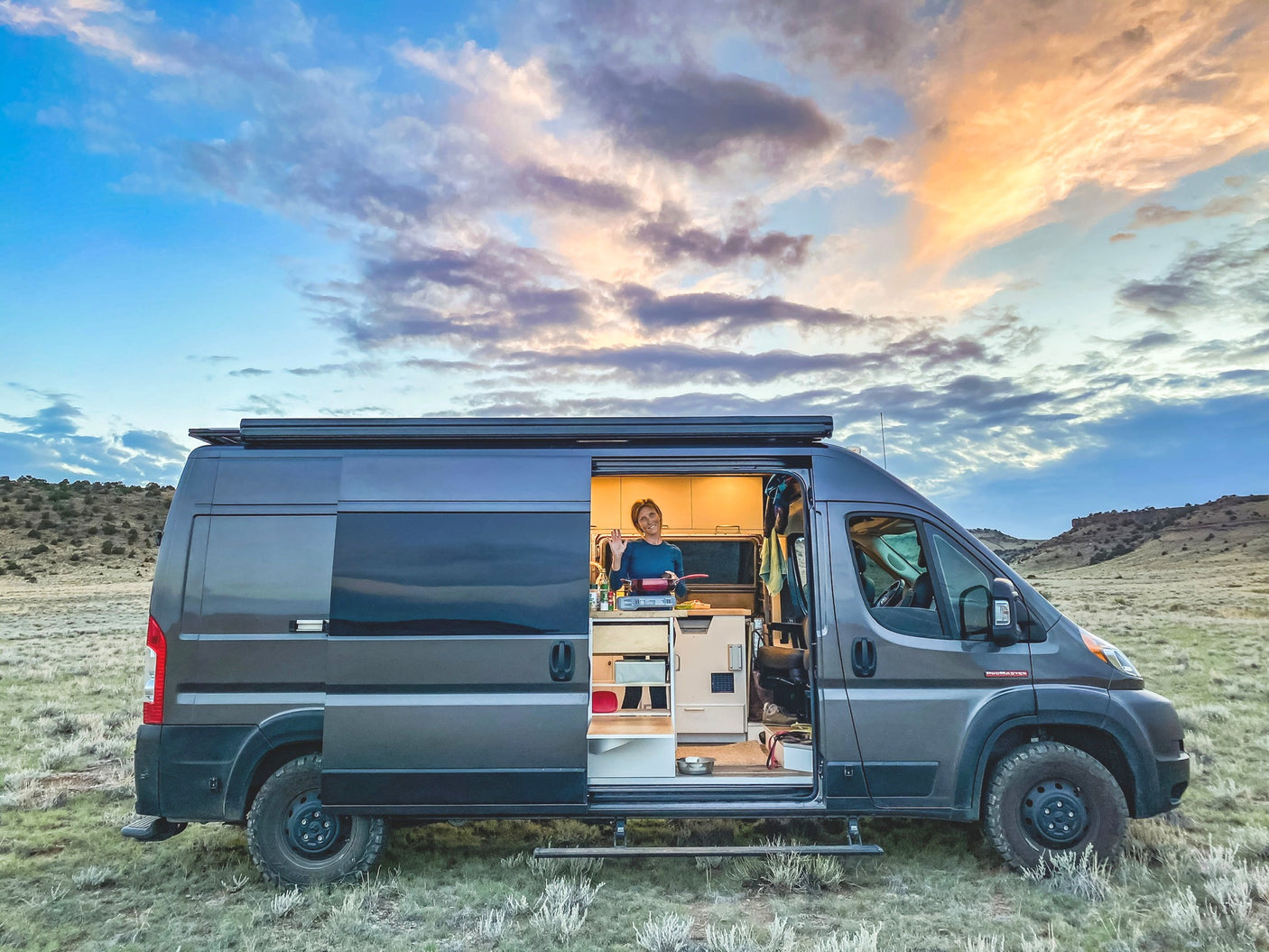Your next adventure starts with an RV rental! A woman cooks inside her custom camper van, set in a vast, open landscape at dusk.