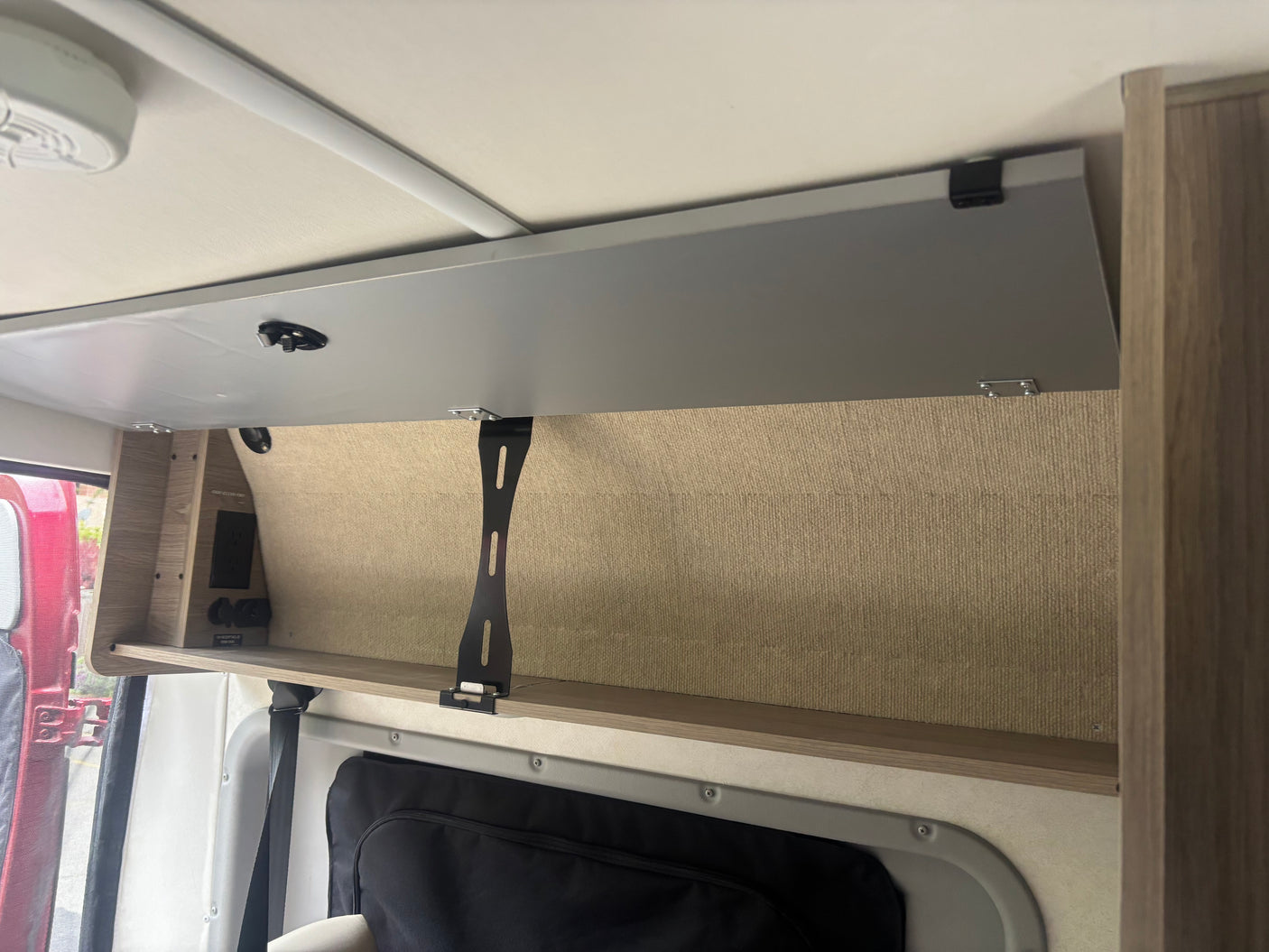 RV rental interior: Open overhead storage, a shelf with AC/USB/12V outlets, and a black support bracket in a camper van.