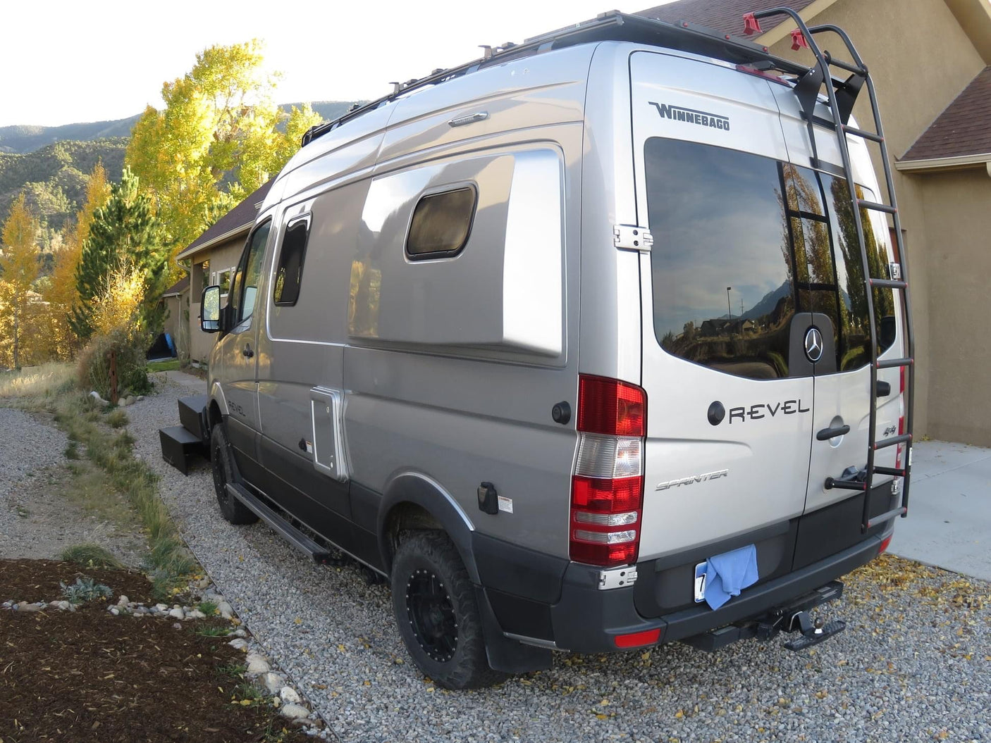 A silver Winnebago Revel 4x4 Mercedes Sprinter camper van on a gravel driveway. Consider this for your rv rental.