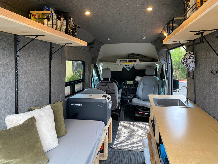 Cozy RV rental camper van interior with bed, Dometic fridge, kitchen sink, wooden shelving, and front seats visible.