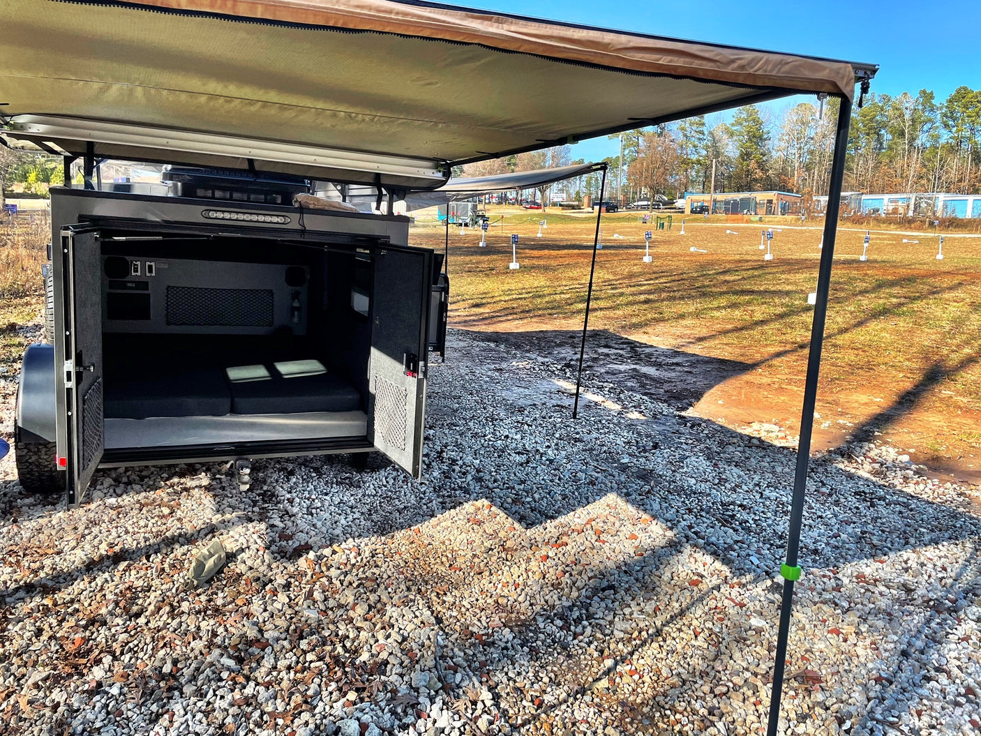 This compact RV rental features an open rear revealing a bed, with an awning extended over a gravel outdoor area.