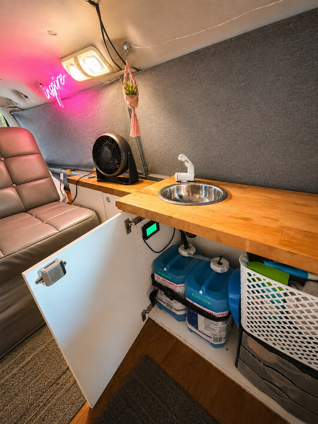 Inside this RV rental: a practical kitchen setup with a sink, hidden water tanks, a fan, & a glowing 'inspire' sign.