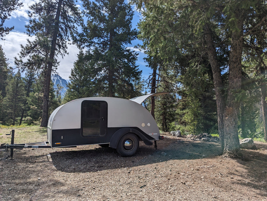 Experience camping! A stylish grey teardrop RV rental parked on a forest campsite with mountain views.