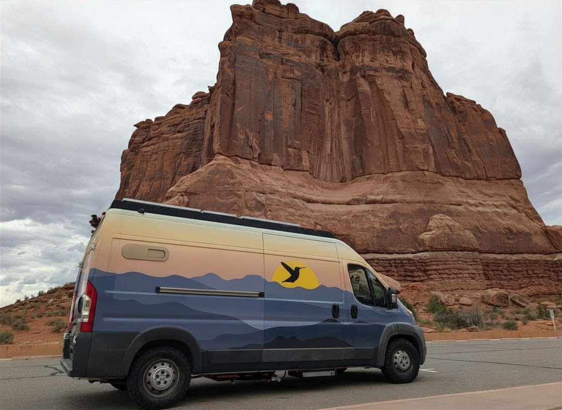 This scenic rv rental shows a custom camper van by towering red rock formations under a cloudy sky. Great for exploring national parks.