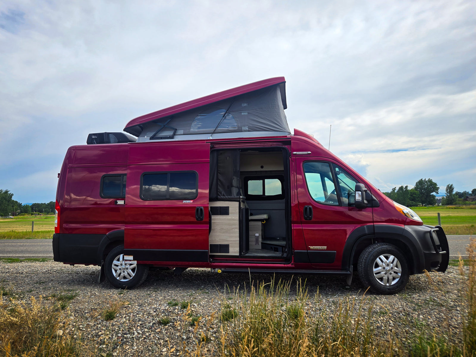This red pop-top RV rental is parked on a gravel shoulder, its open side door revealing the interior against fields and sky.