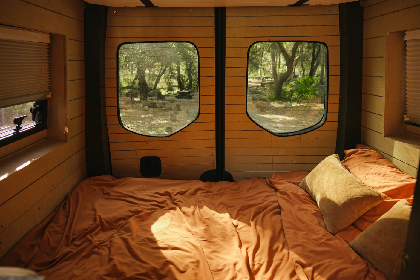 Inside a cozy rv rental, a comfortable bed with orange sheets faces two rear windows offering a tranquil forest view.