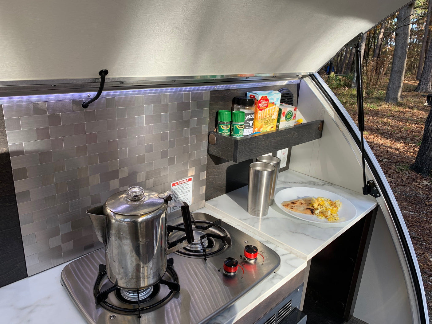 Teardrop RV rental kitchen galley with stove, coffee percolator, and breakfast, ready for outdoor meals.