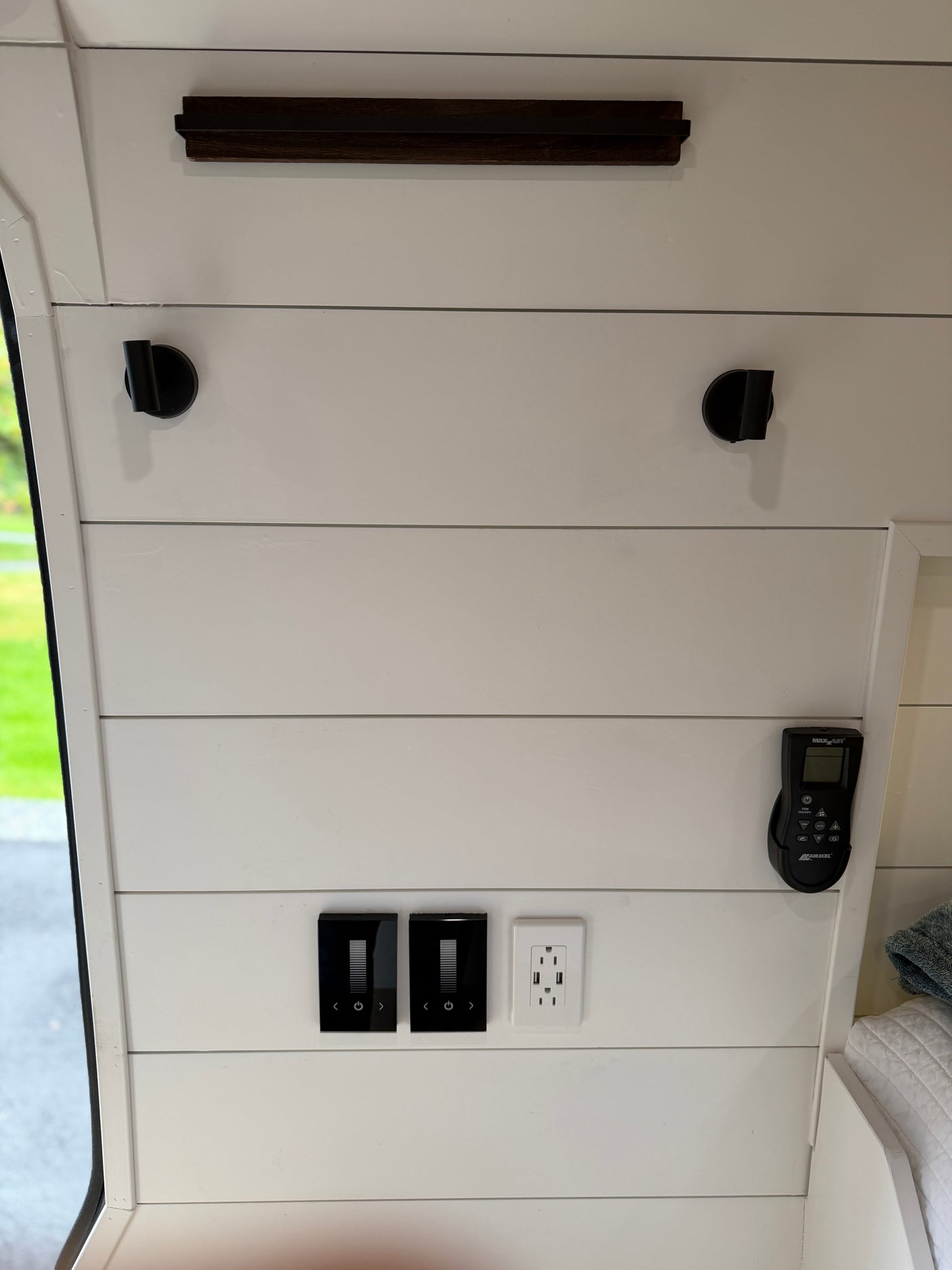 RV rental interior: White shiplap wall features modern light controls, USB outlet, MAXAIR remote, hooks, and a dark shelf.