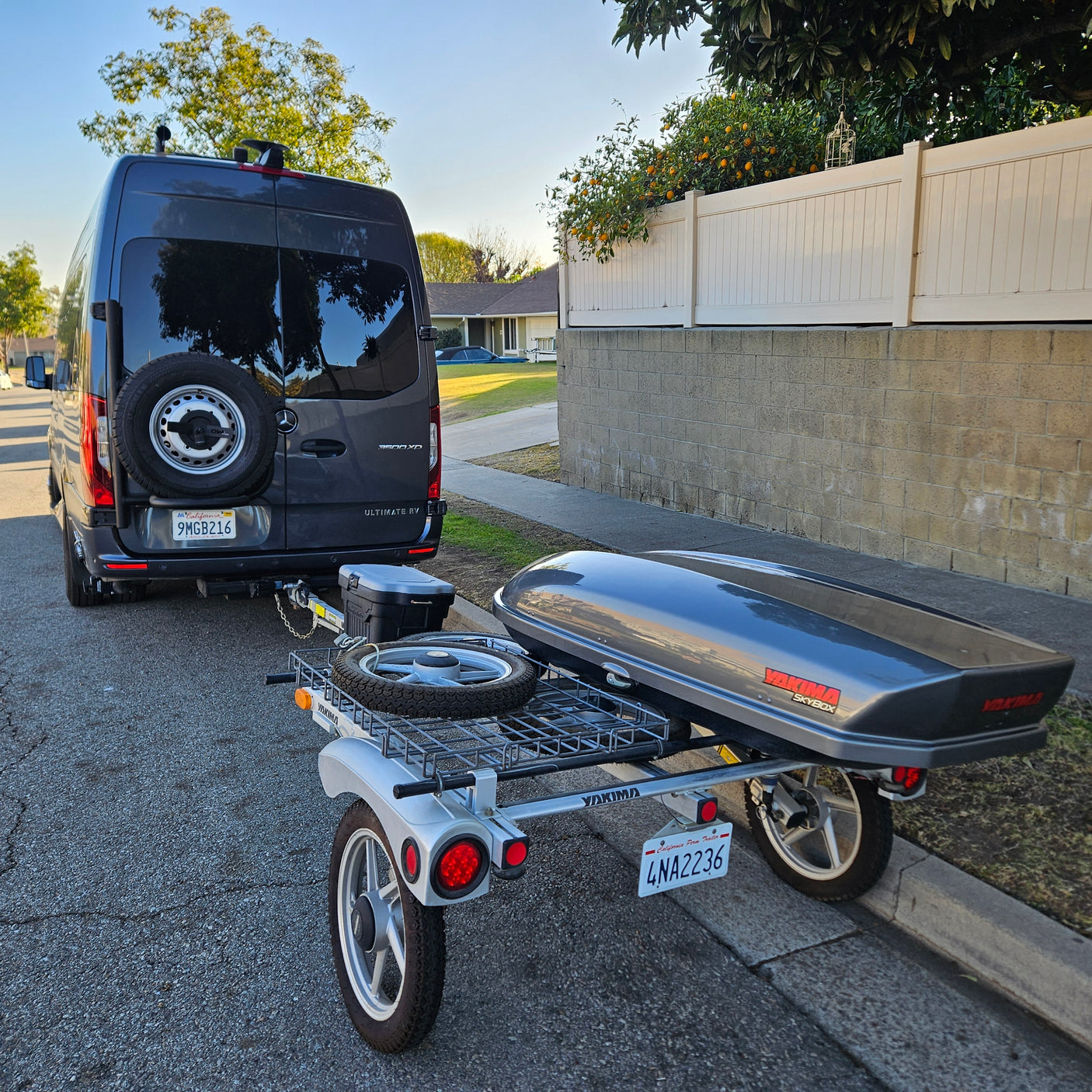 Dark gray Mercedes Sprinter RV rental van towing a compact trailer with a Yakima SkyBox cargo box and a spare tire.