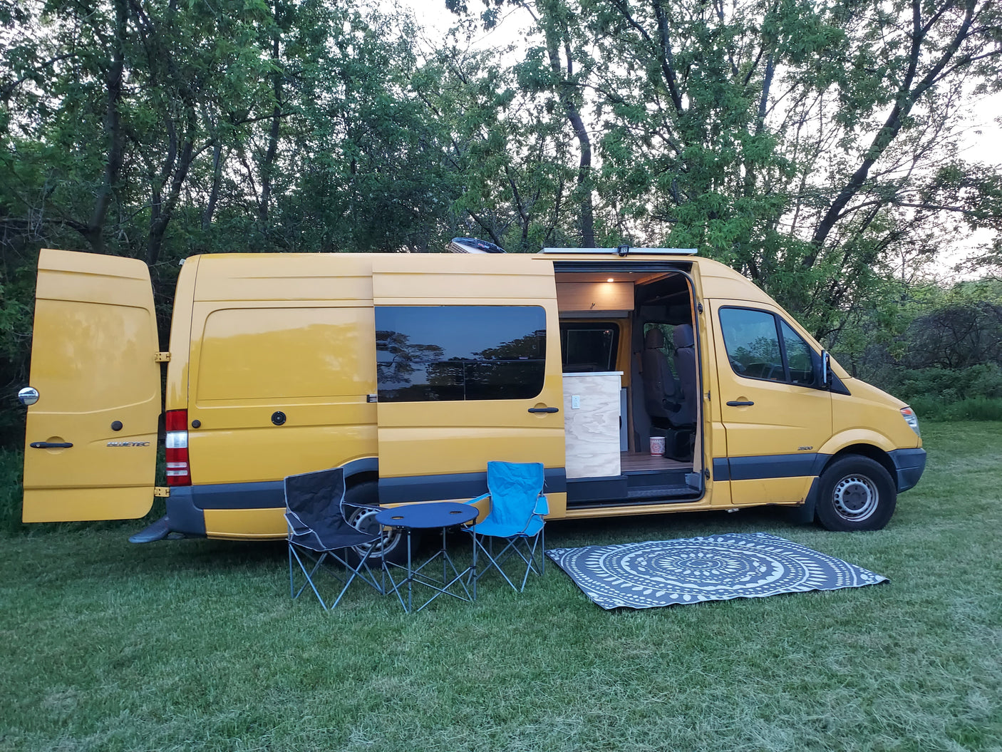 Yellow RV rental camper van with open doors, outdoor chairs, and a mat on grass, set for a relaxing stop.