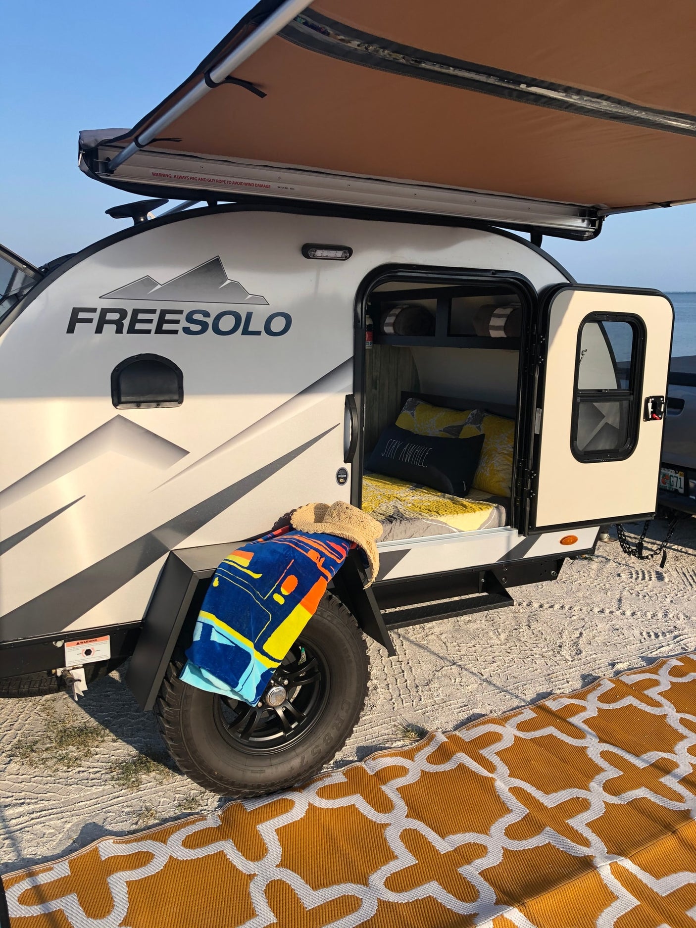 FREESOLO teardrop RV rental set up by the water, awning extended, door open to interior. Perfect for beach camping.
