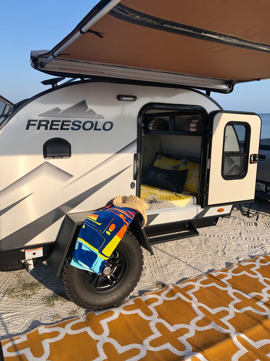 FREESOLO teardrop RV rental set up by the water, awning extended, door open to interior. Perfect for beach camping.