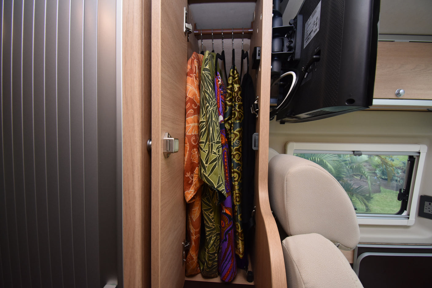 Inside an RV rental: an open wardrobe with colorful clothes, a mounted TV, and a window showing a green outdoor view.