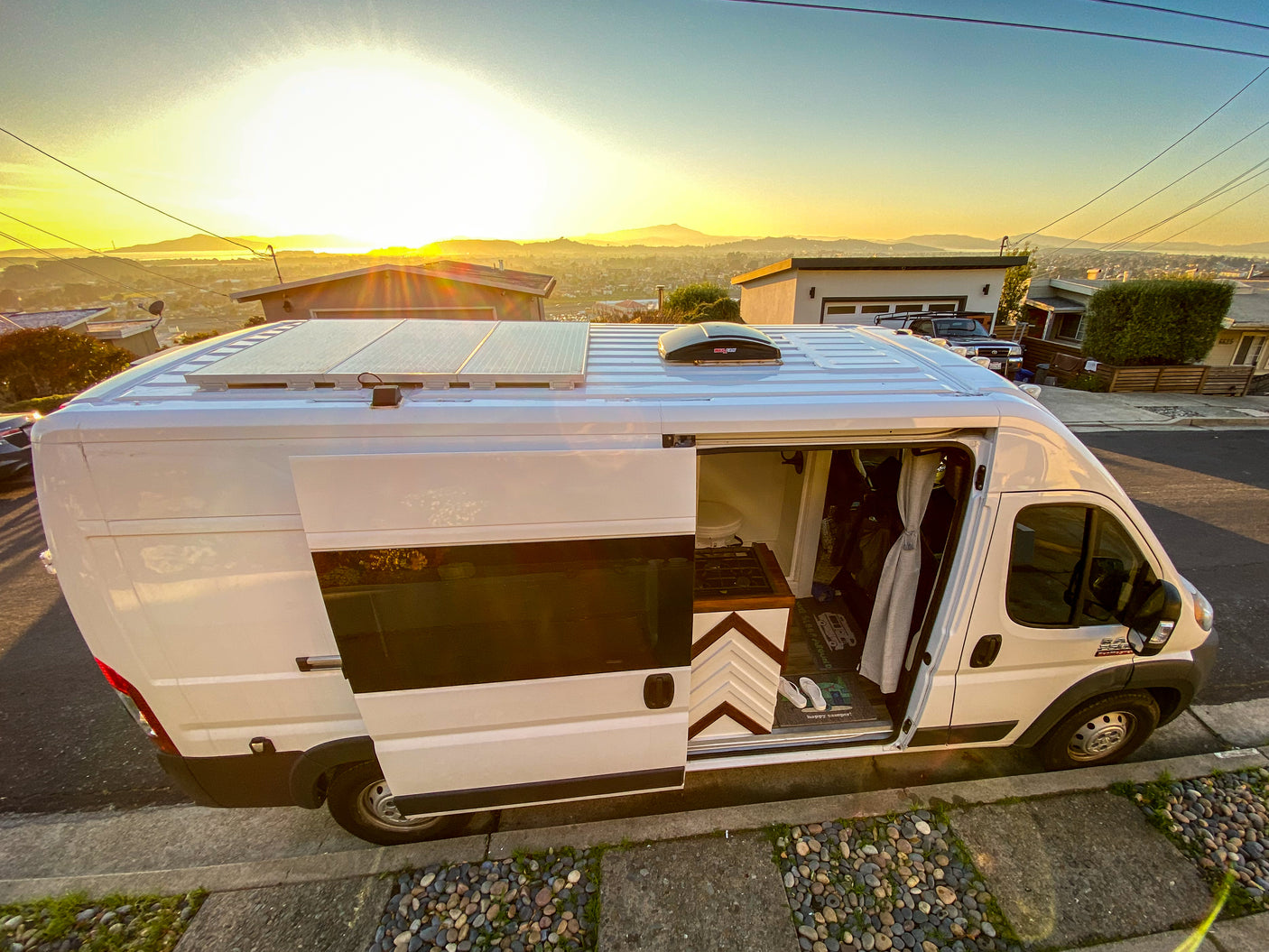 White RV rental camper van parked on a street at sunset. Its open door reveals a kitchen, small toilet, and solar panels on top.