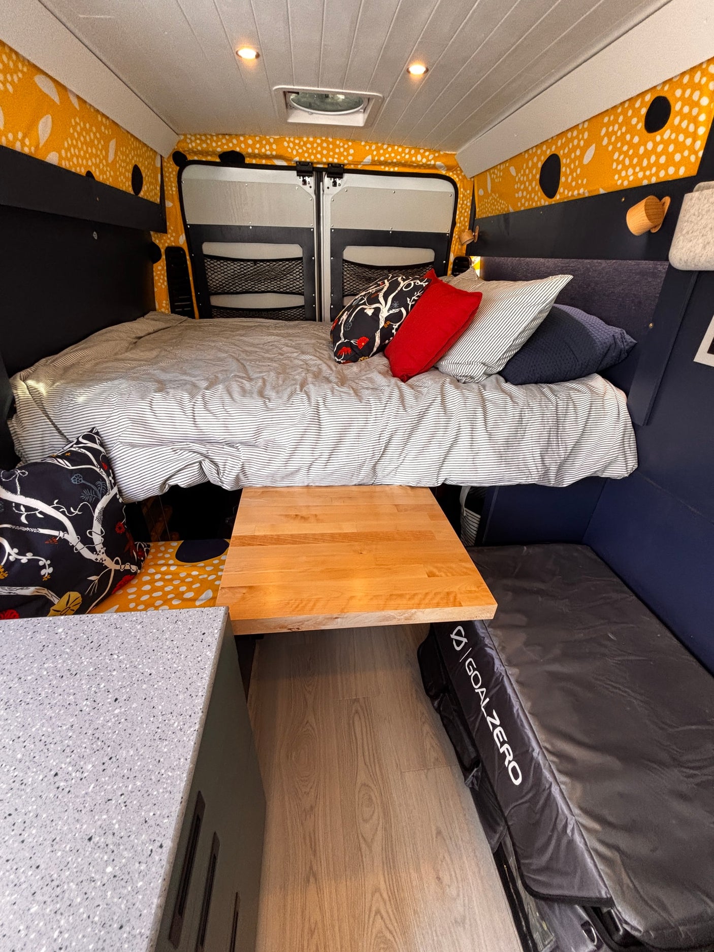RV rental: Camper van interior with a cozy striped bed, wood table, speckled countertop, and unique yellow patterned walls.