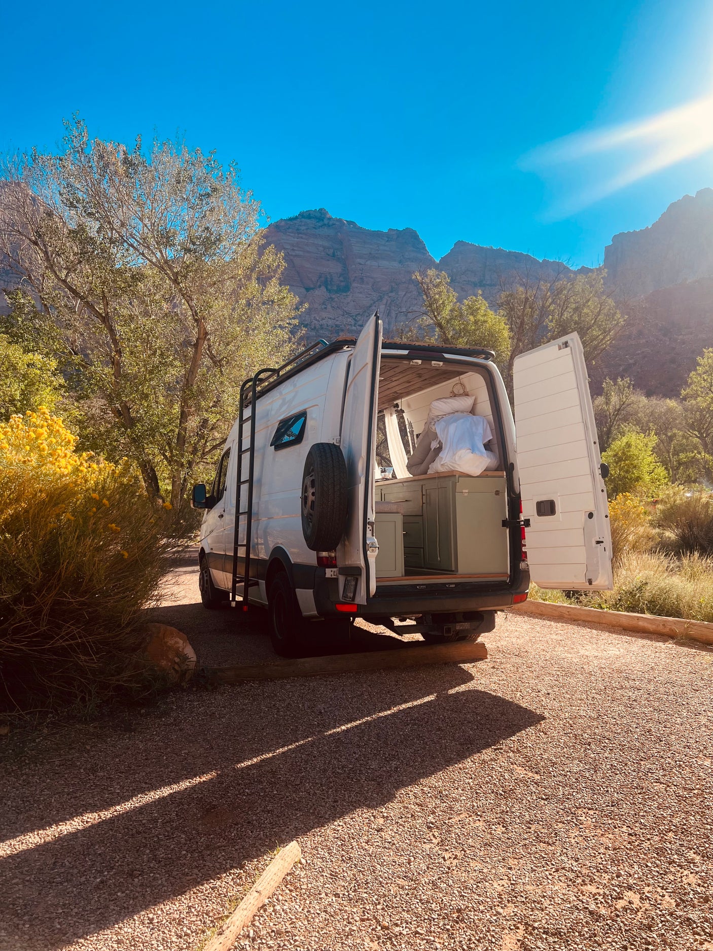 White RV rental van with open back doors, showing interior, parked on gravel amidst trees & mountains under bright sun.
