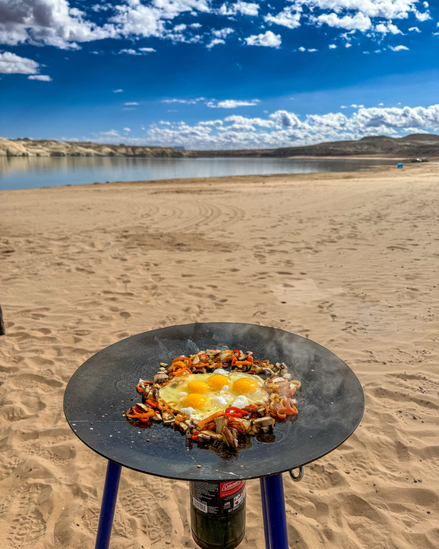 RV rental trip: Outdoor cooking setup with eggs, mushrooms, and peppers on a griddle at a sandy lake beach under blue skies.