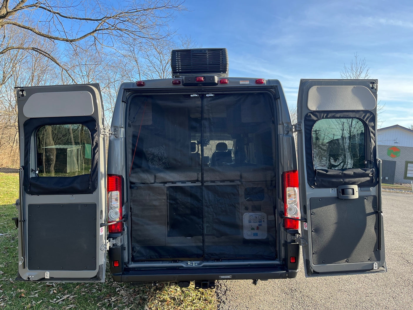 This RV rental van's rear doors are open, revealing a bug screen, interior bed, and a rooftop AC unit under a blue sky.
