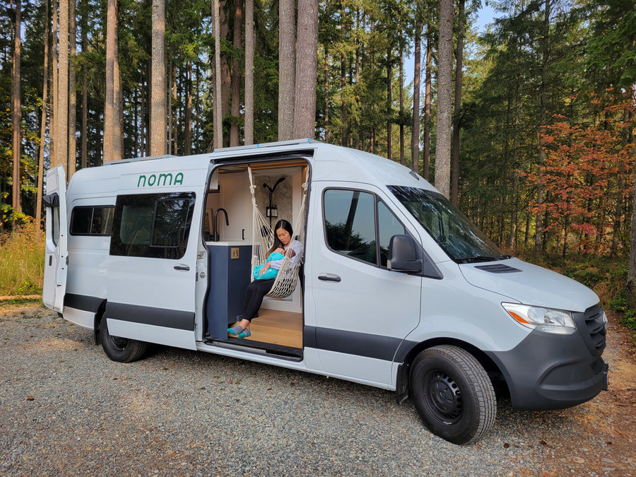 A woman with a baby rests in a hanging chair inside a white NOMA RV rental campervan, parked in a forest.