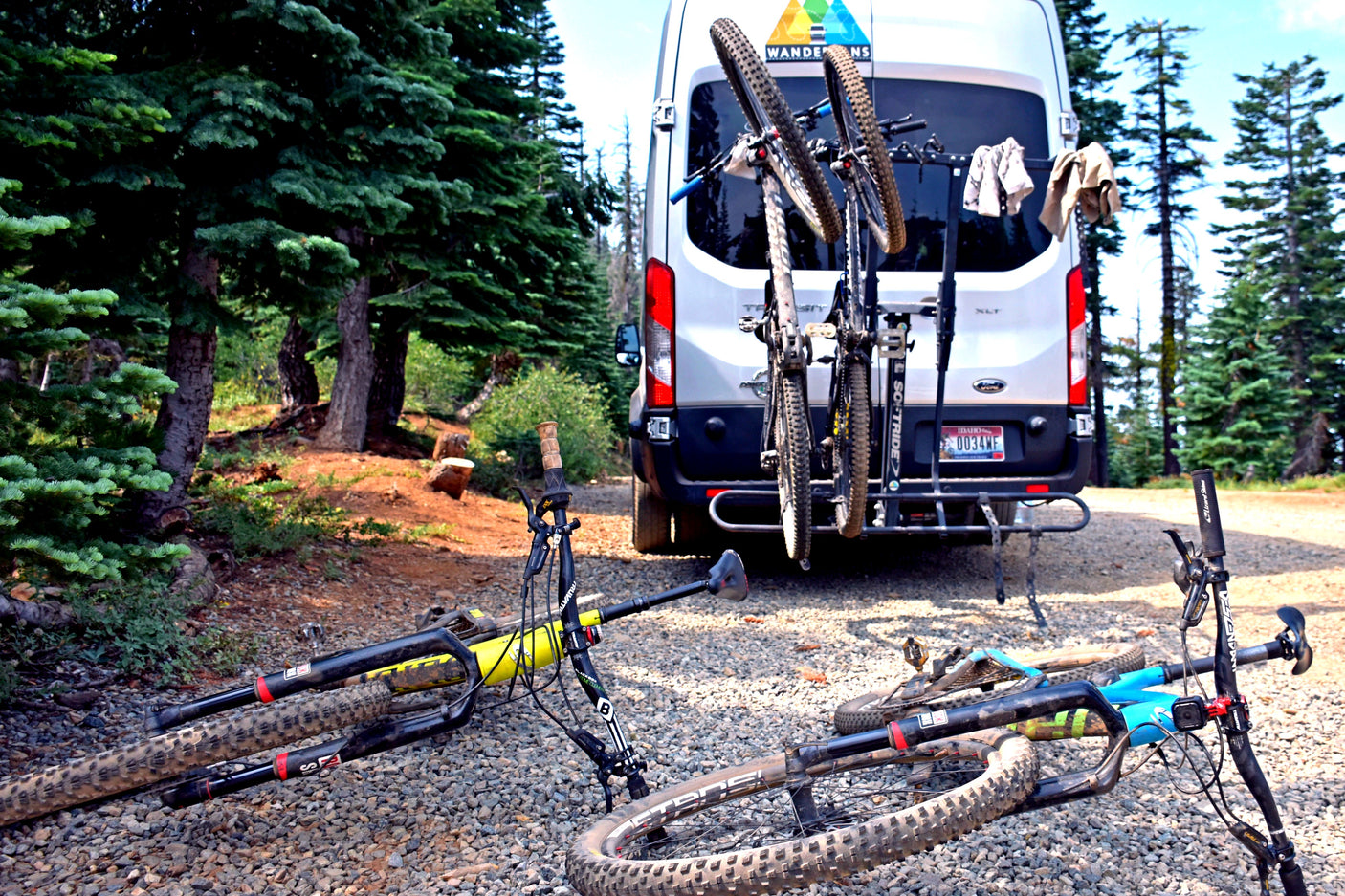 White RV rental van with bikes on a rack and two mountain bikes on the ground, set in a peaceful forest.