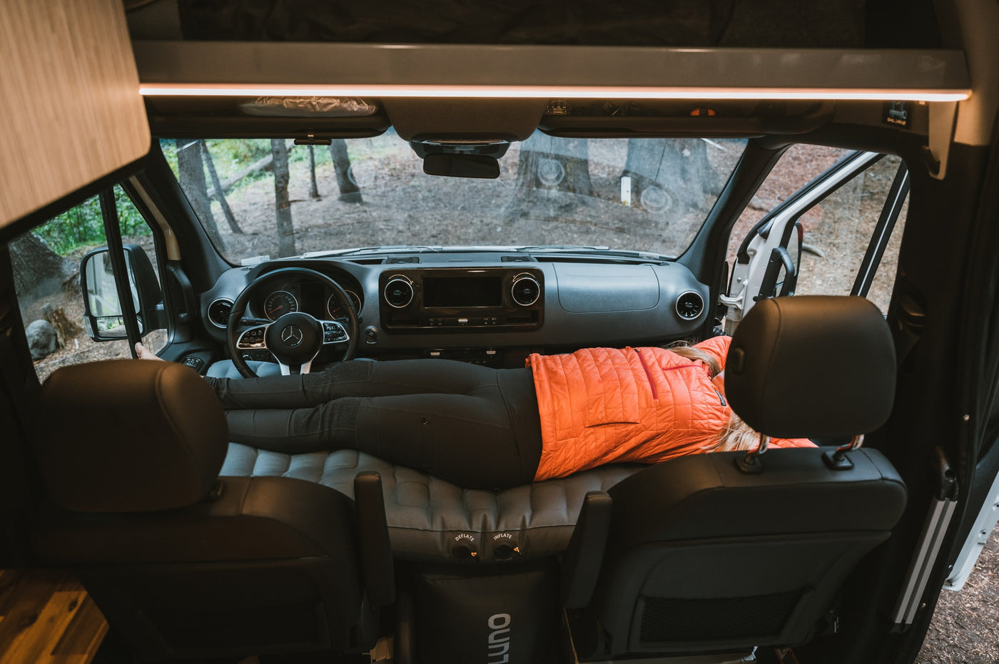 A person in an orange jacket rests on a Luno air mattress inside an RV rental van, making the front cab a cozy sleeping area. — photo 12