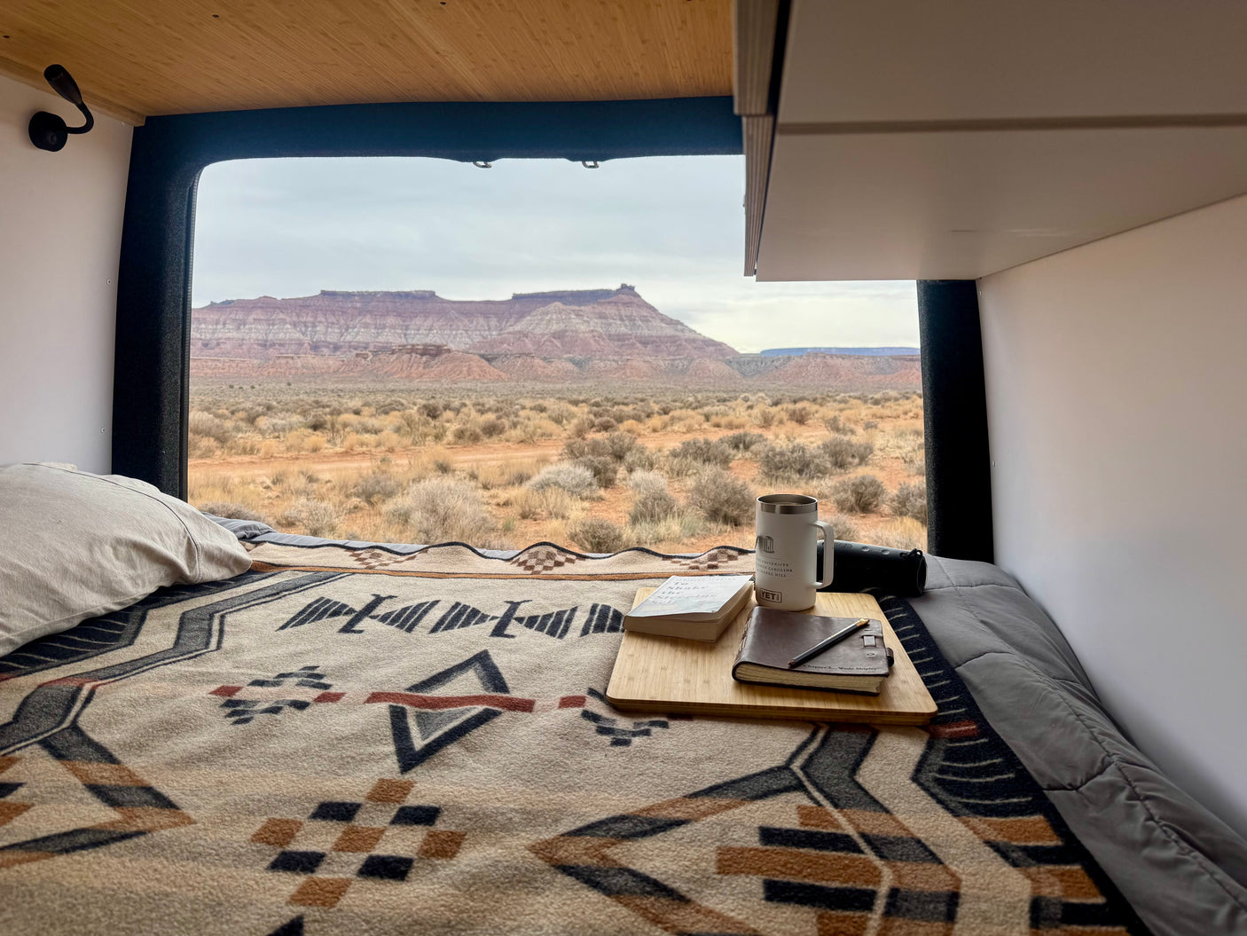Cozy RV rental interior: Bed with patterned blanket, books, and coffee mug, overlooking a vast desert and mountains.
