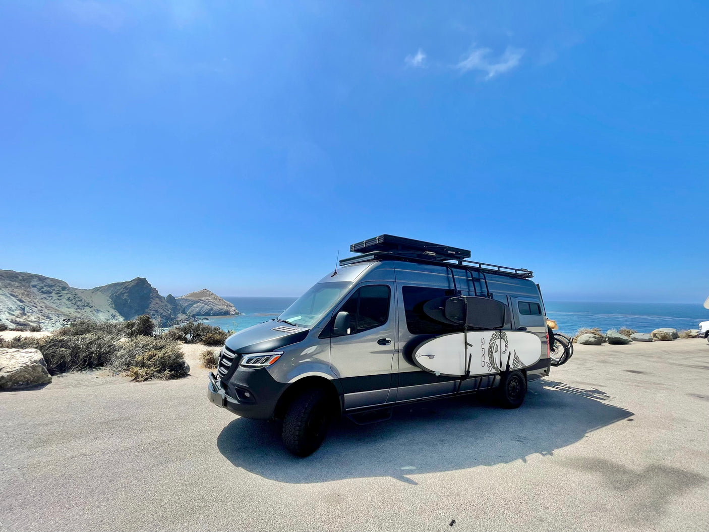 Coastal adventure awaits! Modern rv rental camper van with surfboards & bike overlooking the ocean.