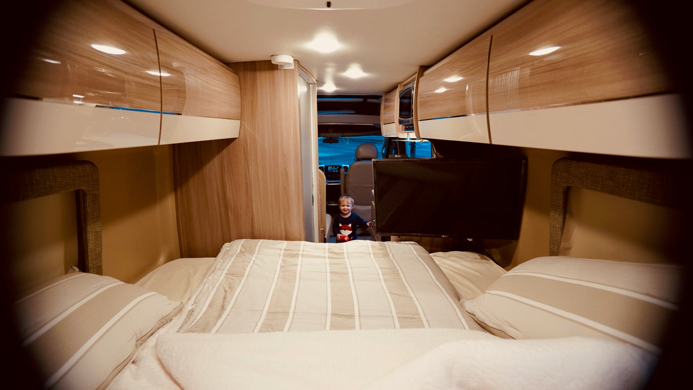 Cozy RV rental interior: A happy child stands near the front, framed by a bed, sleek cabinets, and a mounted TV. Ideal for family road trips.
