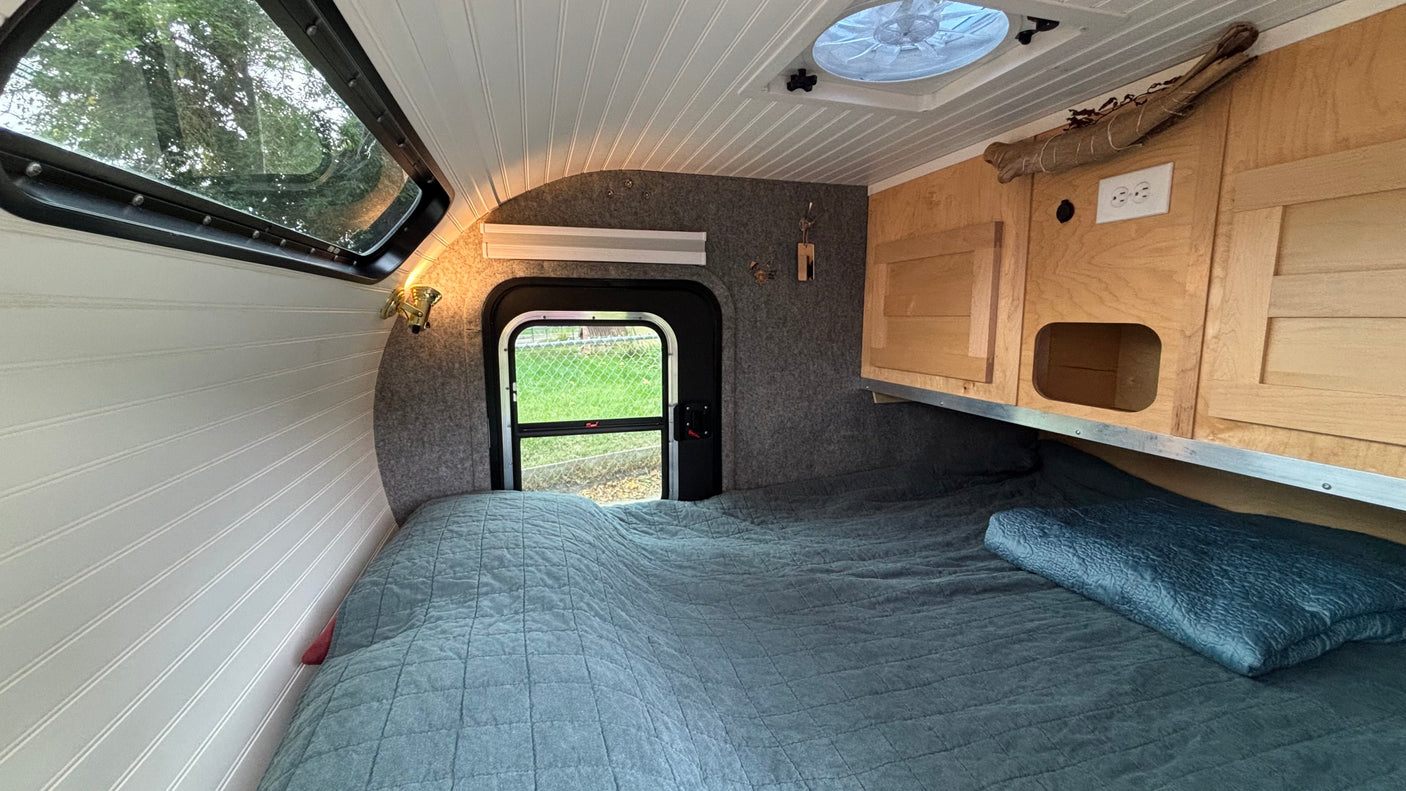 Inside a cozy RV rental: quilted bed, wood cabinets, ceiling vent, and window create a comfortable camper interior.