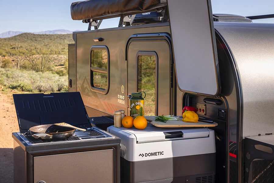 Enjoy outdoor cooking with this RV rental setup: portable stove, Dometic cooler, and fresh ingredients next to an adventure trailer.