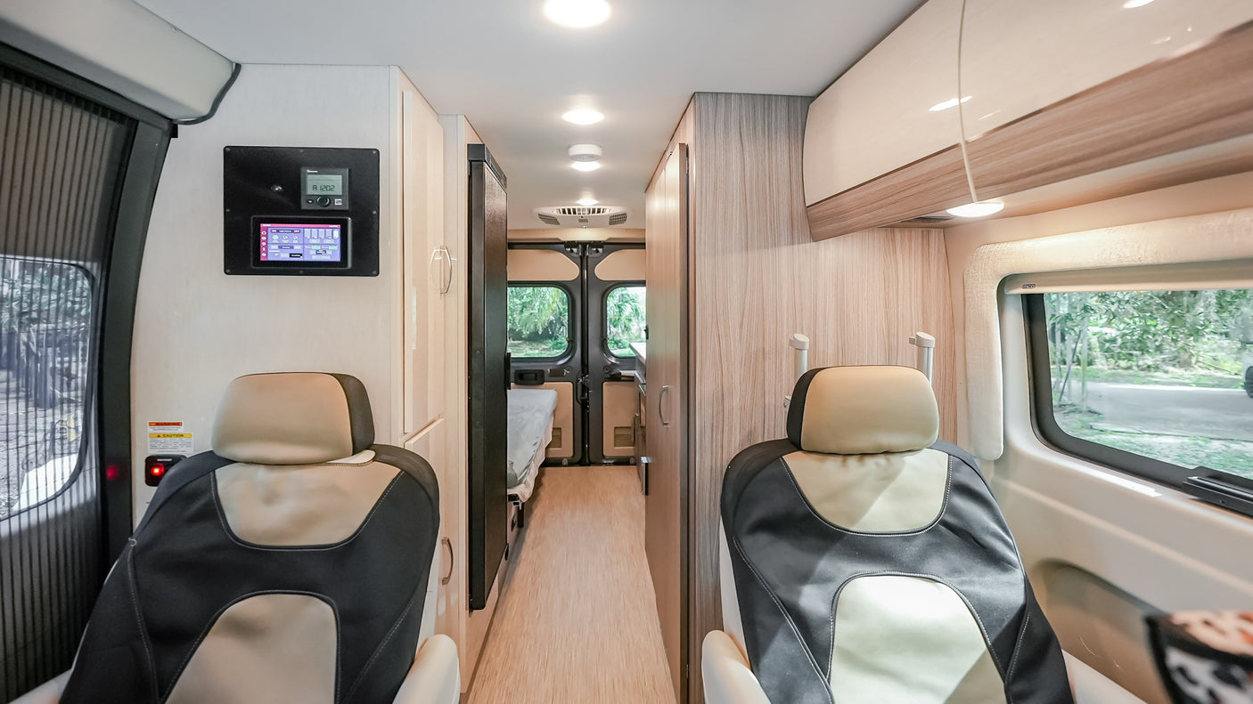Modern RV rental interior: front seats, wall controls, bright hallway leading to kitchen, and rear bed.