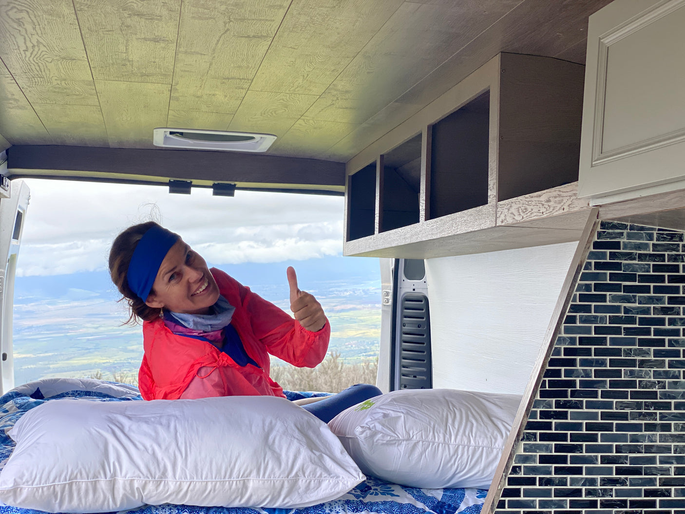 Smiling woman gives thumbs up from cozy RV rental bed, enjoying the stunning scenic view. Perfect for your next van life adventure!