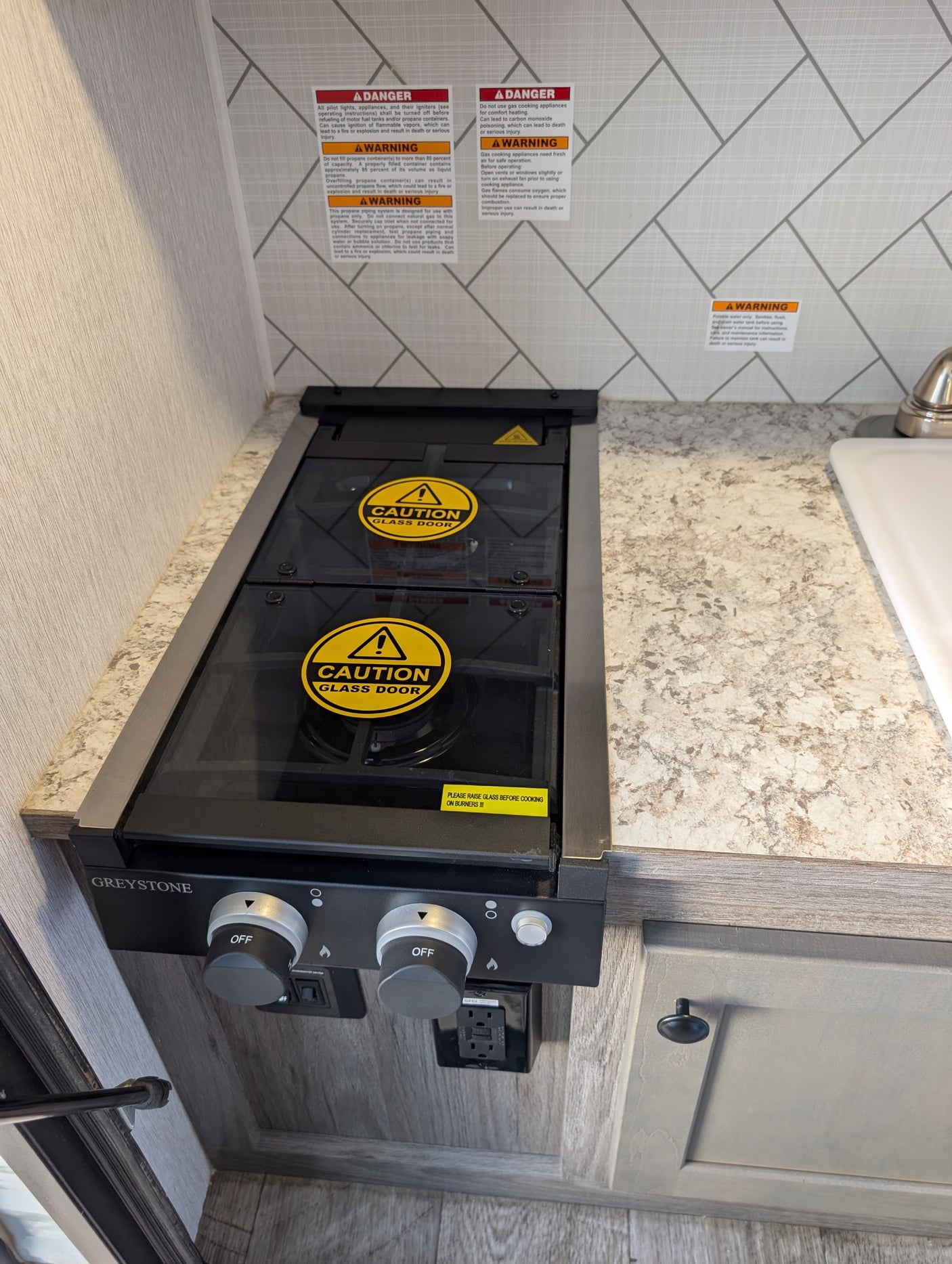 RV rental compact kitchen with a Greystone gas stove, glass cover, safety warnings, and a GFI power outlet.