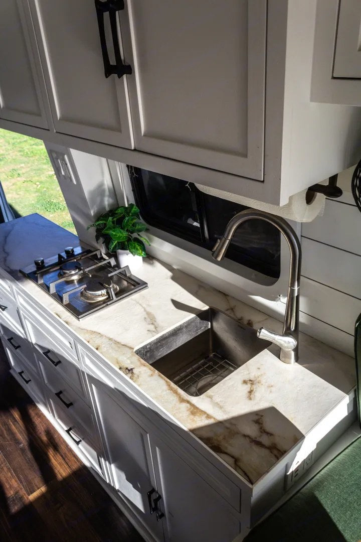 Functional RV rental kitchen: white shaker cabinets, marble-pattern counter, sink, and two-burner stove. Sunlit interior.