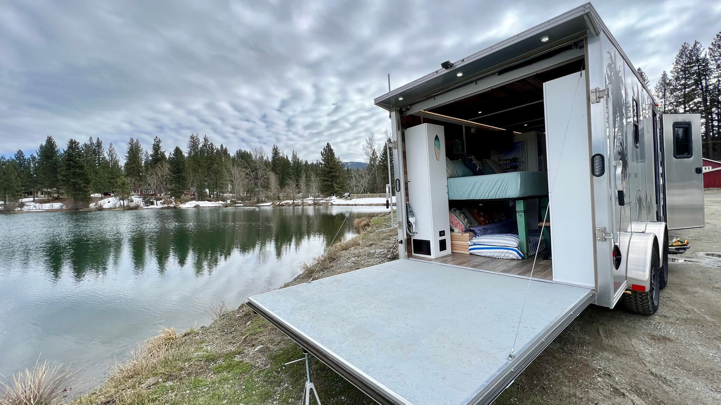 This RV rental offers lakeside camping! A travel trailer with its back open, revealing bunk beds by a calm lake.