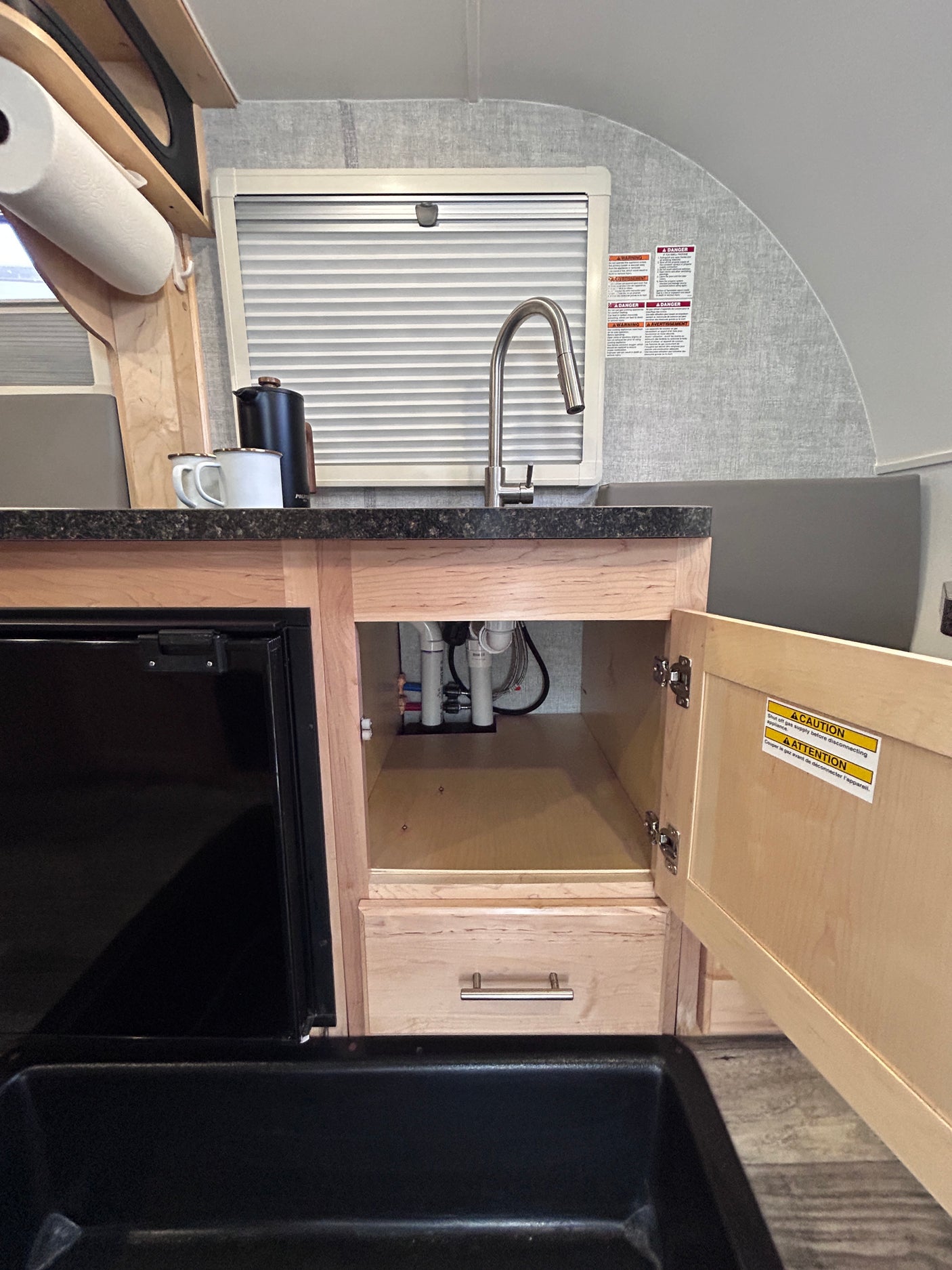 A compact RV rental kitchen features a black sink, faucet, small fridge, and open under-sink cabinet revealing plumbing.