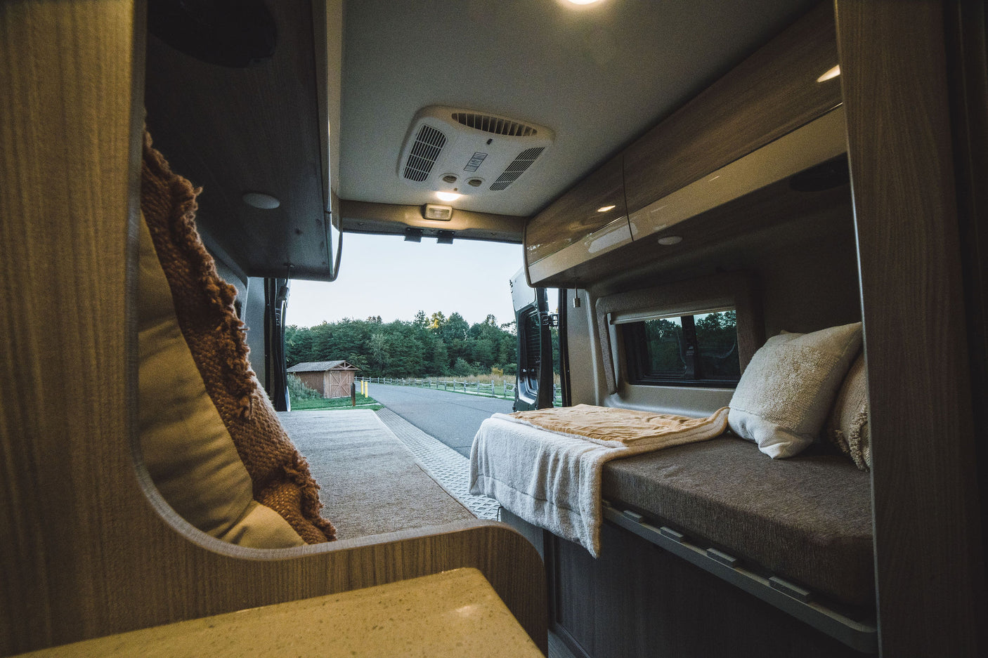 Cozy RV rental interior with bed, pillows, AC, and open rear doors revealing a scenic road and forest.