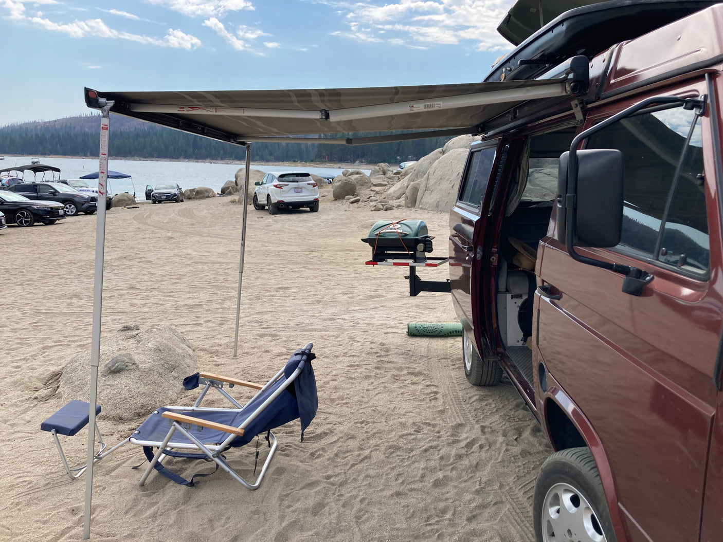 Enjoying the beach! A maroon RV rental camper van with an awning, chairs, and grill setup on a sandy lake shore.